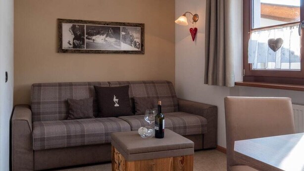 Una sala de estar con un sofá, una mesa de centro de madera con una botella y un vaso, un cuadro enmarcado en la pared y una ventana con cortinas.