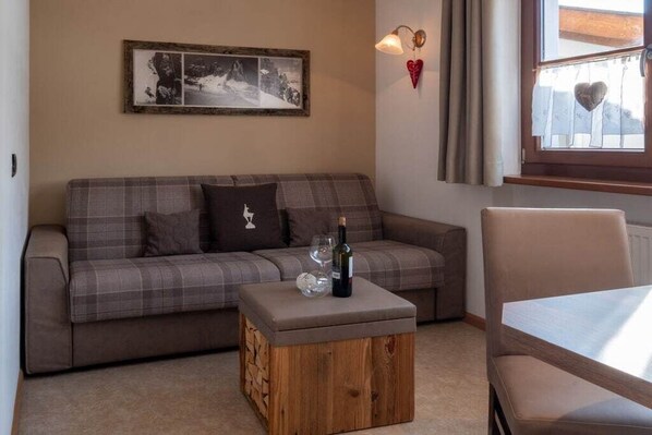 A living room with a sofa, a wooden coffee table with a bottle and glass, a framed picture on the wall, and a window with curtains.