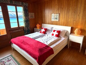 A bedroom with a bed, a nightstand, a lamp, and a window with a view of a lake and mountains. - Concierge, Lake Thun/Alps Scenic Views by SwissHut (Sigriswil)