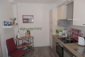 Een compacte keuken met een magnetron, koffiezetapparaat en een klein eethoekje met een stoel en een plank met keukengerei.