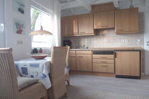 A kitchen with wooden cabinets, a dining area with wicker chairs, and a window providing natural light. - Holiday home with 2 bedrooms (Varel - Dangast)