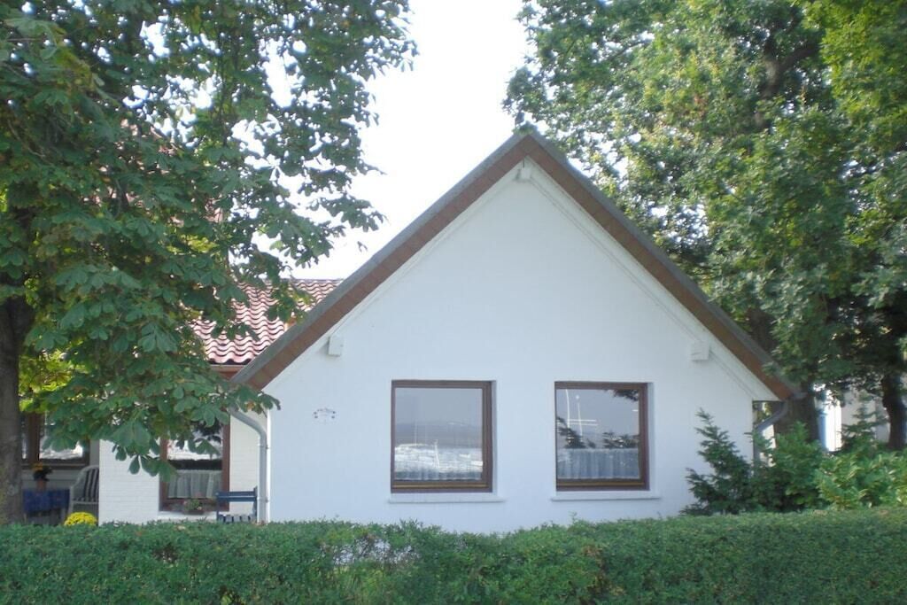 A white house with a gabled roof, two windows with brown frames, and a small porch with a bench.