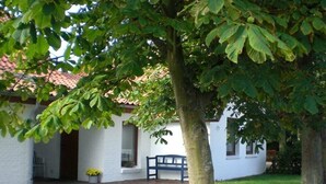 A white building with a red-tiled roof, a bench, and a tree in the foreground. - Holiday home with 2 bedrooms (Varel - Dangast)