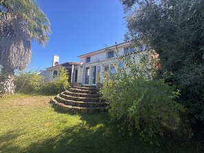 A two-story building with a stone staircase leading to the entrance, surrounded by lush greenery and a clear blue sky. - Country house near the city, peace and quiet, nature, a pool, and a spacious garden. (Bucelas)