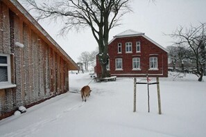 A snow-covered outdoor area with a wooden building and a brick house in the background. - Holiday home with 2 bedrooms (Forlitz-Blaukirchen)