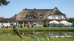 Un grand bâtiment avec un toit de chaume, un étang au premier plan et des espaces aménagés pour s’asseoir à l’extérieur.