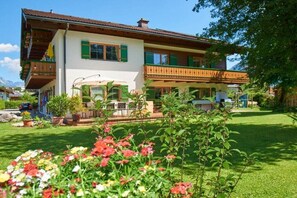 Ein zweistöckiges Gebäude mit einem Balkon und einem Garten voller Blumen, unter einem klaren Himmel.