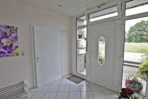 Quarto com porta branca, um banco e vista para uma parede de tijolos e vegetação lá fora.