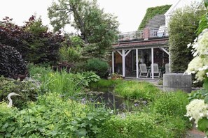 Ein Garten mit Teich, eine überdachte Terrasse mit Stühlen und ein Backsteinhaus mit Balkon.