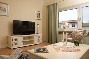 A living room with a flat-screen TV on a white cabinet, a table with a magazine and candles, and a wicker chair. - Holiday apartment with 1 bedroom (Grünendeich)