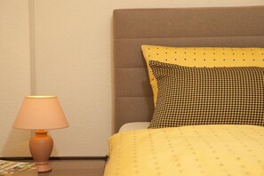 Ein Bett mit einem karierten Kopfkissen und einer gelben Decke, eine Nachttischlampe sowie ein Buch auf dem Nachttisch.