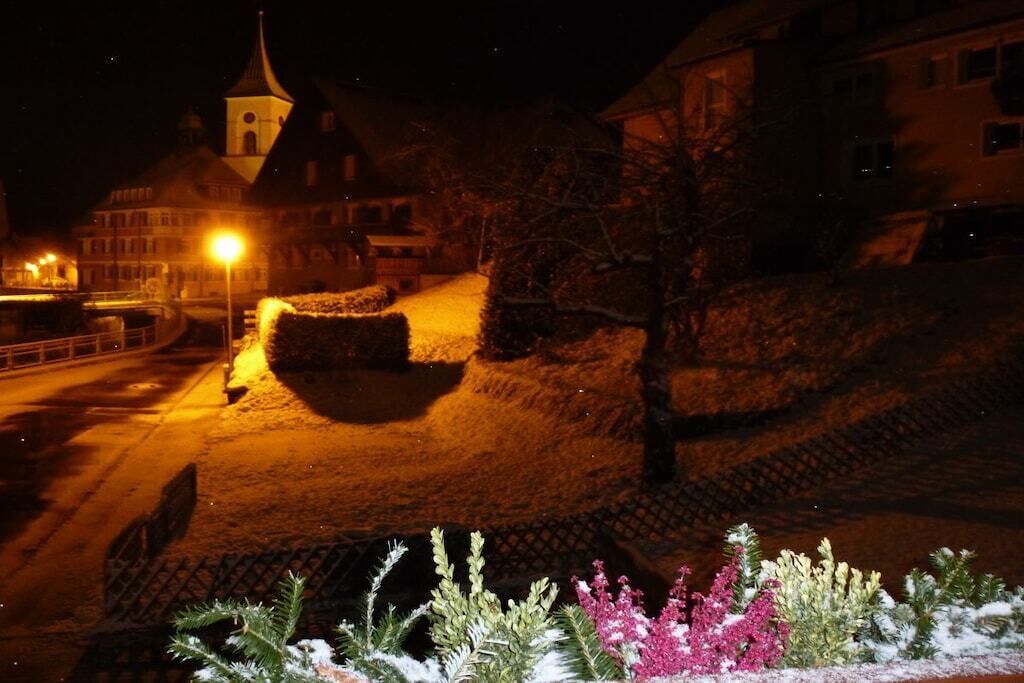Una vista notturna di una cittadina con terreno innevato, una chiesa con una torre e una strada illuminata da lampioni.
