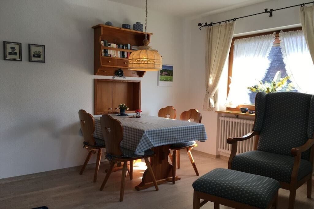 Un comedor con una mesa dispuesta para cuatro, sillas de madera, una lámpara colgante, una estantería y un ventanal con cortinas.