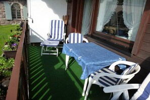 An outdoor patio with a table and two striped chairs, a small garden, and a building with a window. - Holiday apartment with 2 bedrooms (Lenzkirch)