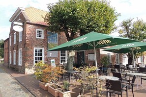A brick building with outdoor seating and umbrellas, a tree, and a signboard. - Holiday apartment with 2 bedrooms (Krummhörn-Greetsiel)