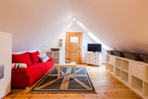A cozy attic bedroom with a red sofa, a wooden door, a television, and a shelf with baskets. - Holiday apartment with 2 bedrooms (Krummhörn-Greetsiel)