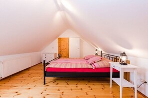 Une chambre avec un plafond en pente, un lit avec une literie rouge, une table de chevet blanche surmontée d’une lanterne, et un plancher en bois.