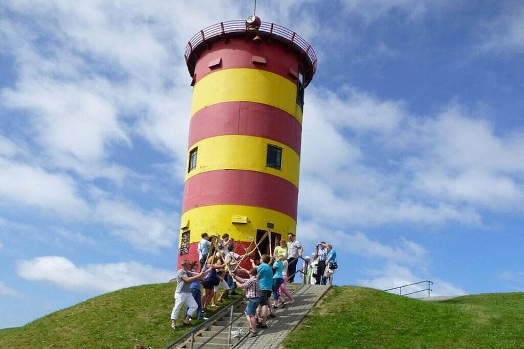 Ein Leuchtturm mit rot-gelben Streifen an der Außenwand, eine Treppe, die zu ihm hinaufführt, und eine Gruppe von Menschen, die die Treppe hinaufsteigt.
