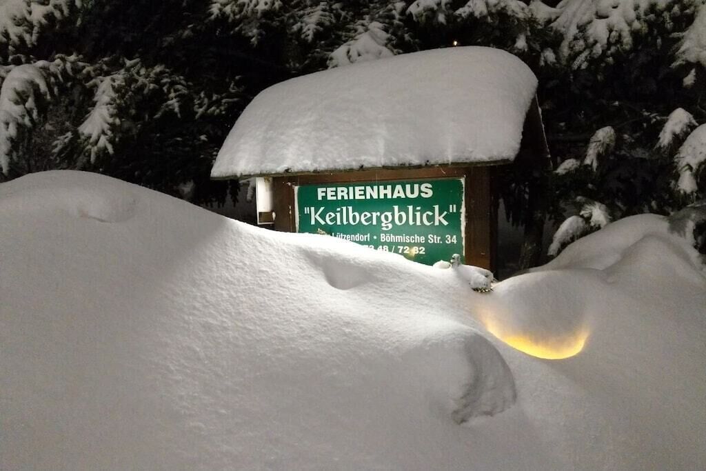 Un cartel cubierto de nieve con la inscripción 'FERIENHAUS Keilbergblick' y un edificio parcialmente visible al fondo.