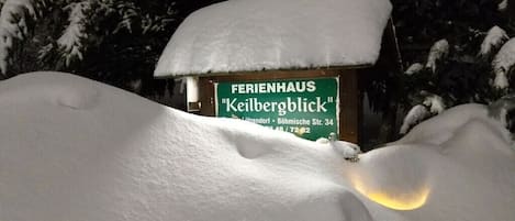 Un cartel cubierto de nieve con la inscripción 'FERIENHAUS Keilbergblick' y un edificio parcialmente visible al fondo.