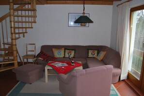 Ein Wohnzimmer mit einer Holztreppe, einem Sofa mit bunten Kissen, einem Tisch mit rotem Tischläufer und einem Fenster mit Vorhängen.