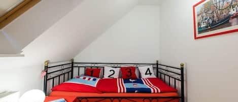 A small, cozy room with a sloped ceiling, a single bed with numbered pillows, a red table with a lamp, and a framed picture on the wall.