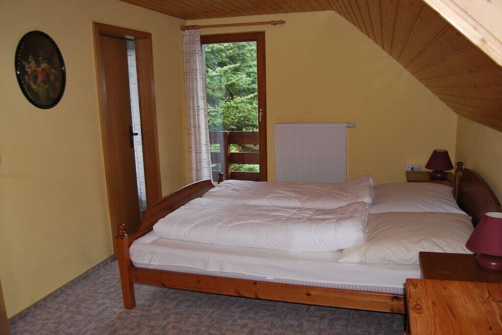 Une chambre avec un lit en bois, une fenêtre donnant sur un paysage verdoyant, un radiateur et une table de chevet avec une lampe.