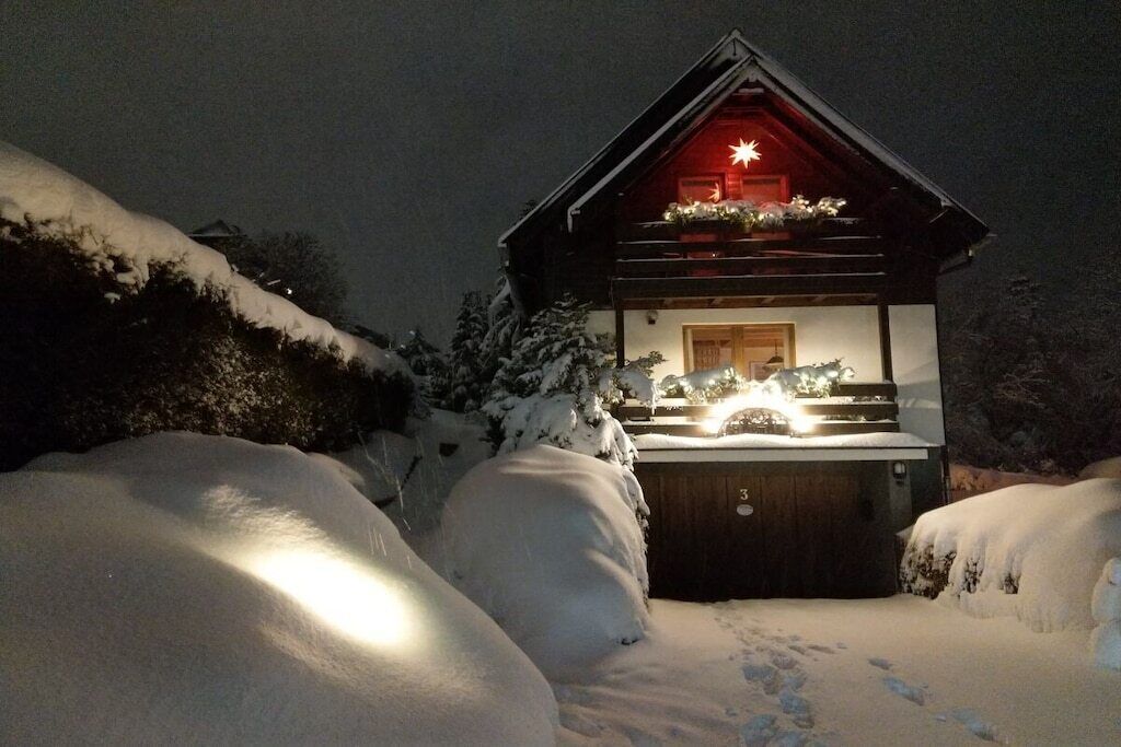 Ein mit Schnee bedecktes Blockhaus mit beleuchtetem Innenraum, Holzfassade und einer verschneiten Auffahrt.