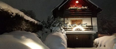 A snow-covered cabin with a lit interior, a wooden exterior, and a snow-covered driveway.