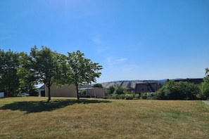 Ein grünes Feld mit einem Baum, einem Gebäude und einer hügeligen Landschaft im Hintergrund.