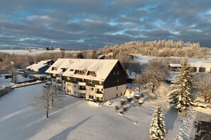 Ein mehrstöckiges Gebäude mit flachem Dach, umgeben von verschneitem Gelände und Bäumen, was eine Winterstimmung vermittelt.