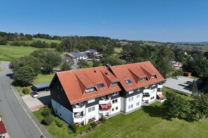Un immeuble à plusieurs étages, surmonté d’un toit recouvert de tuiles rouges, entouré de verdure et baigné d’un ciel dégagé.