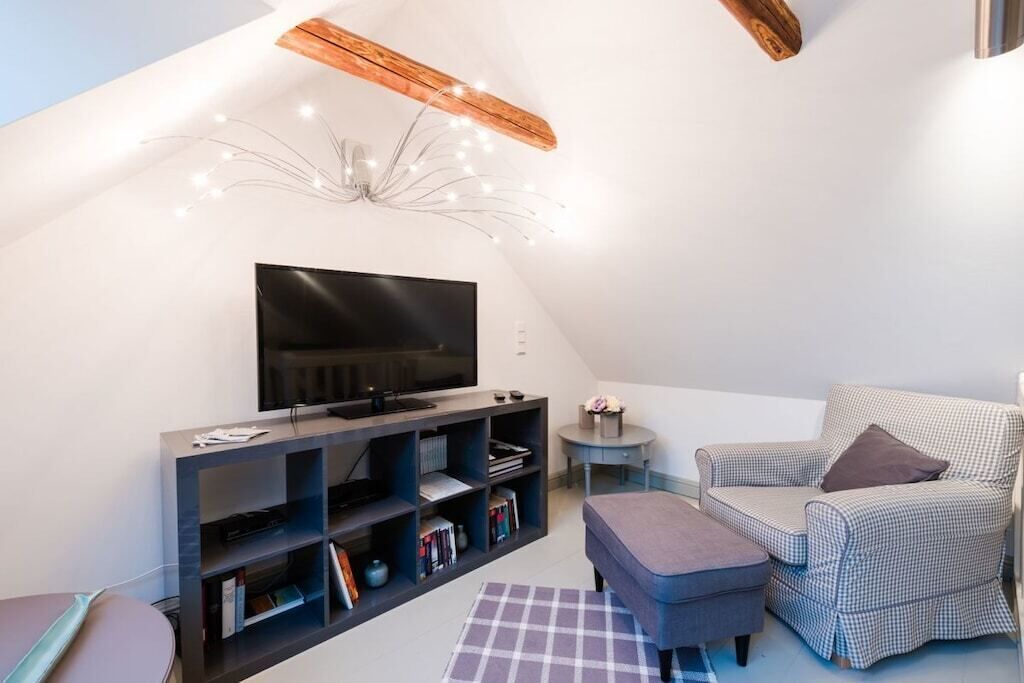 A modern living room with a flat-screen TV, a dark entertainment unit, a checkered armchair, a small side table with a vase, and a grey ottoman.