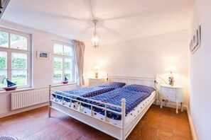 A bedroom with a bed, two bedside tables, a window with curtains, a radiator, and a chandelier. - Holiday home with 2 bedrooms (Krummhörn)