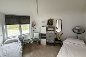 A bedroom with two beds, a window with a view, a bedside table with a lamp, a mirror, and a fan. - Holiday home with 2 bedrooms (Julianadorp)