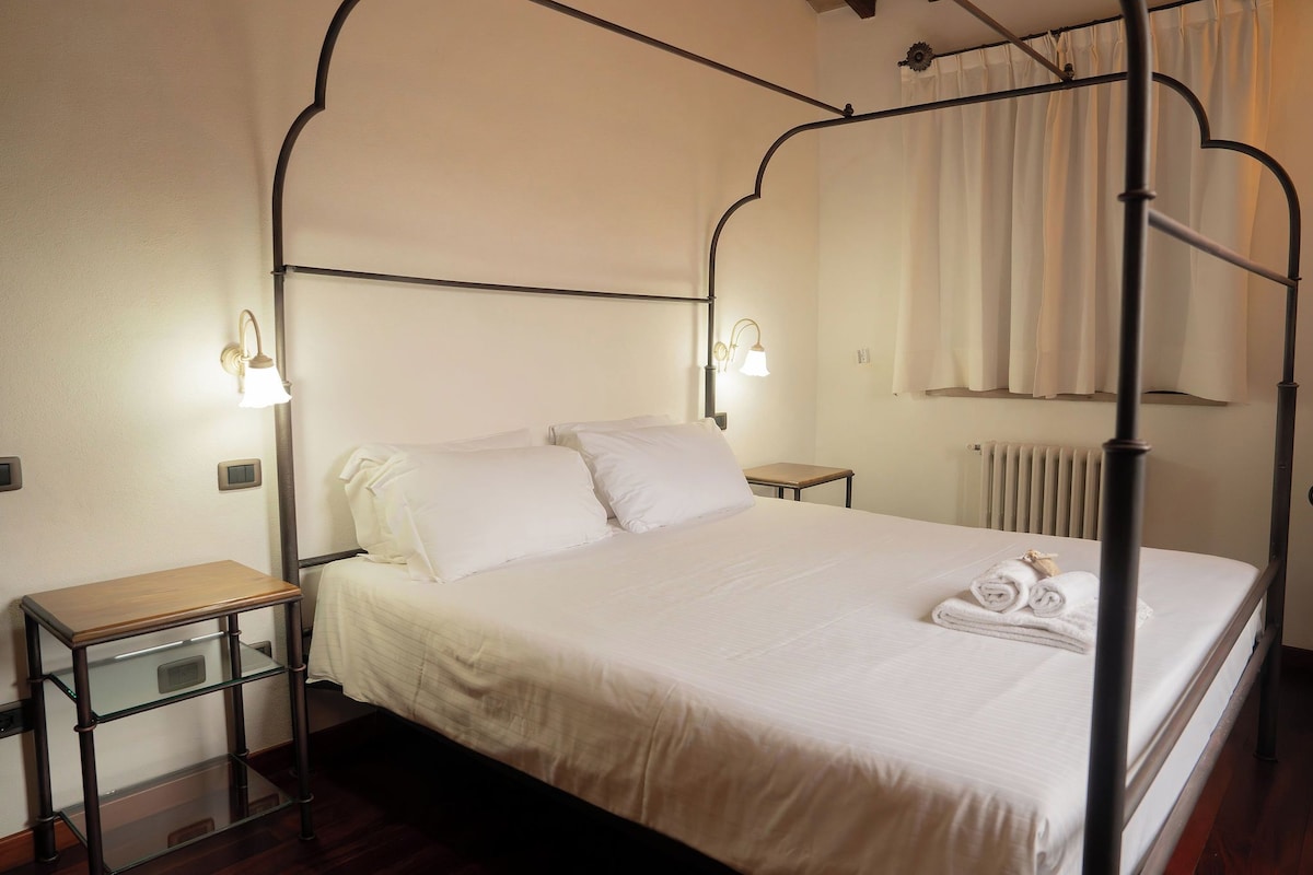 A four-poster bed with a white bedspread and pillows, a bedside table with a lamp, and a radiator in the background.