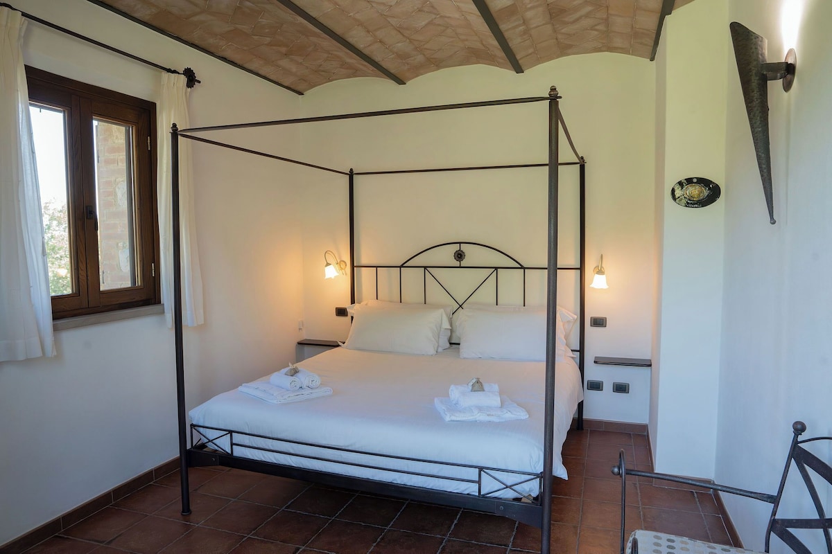 A four-poster bed with white linens, two bedside tables, a window with curtains, and a wall-mounted light fixture.