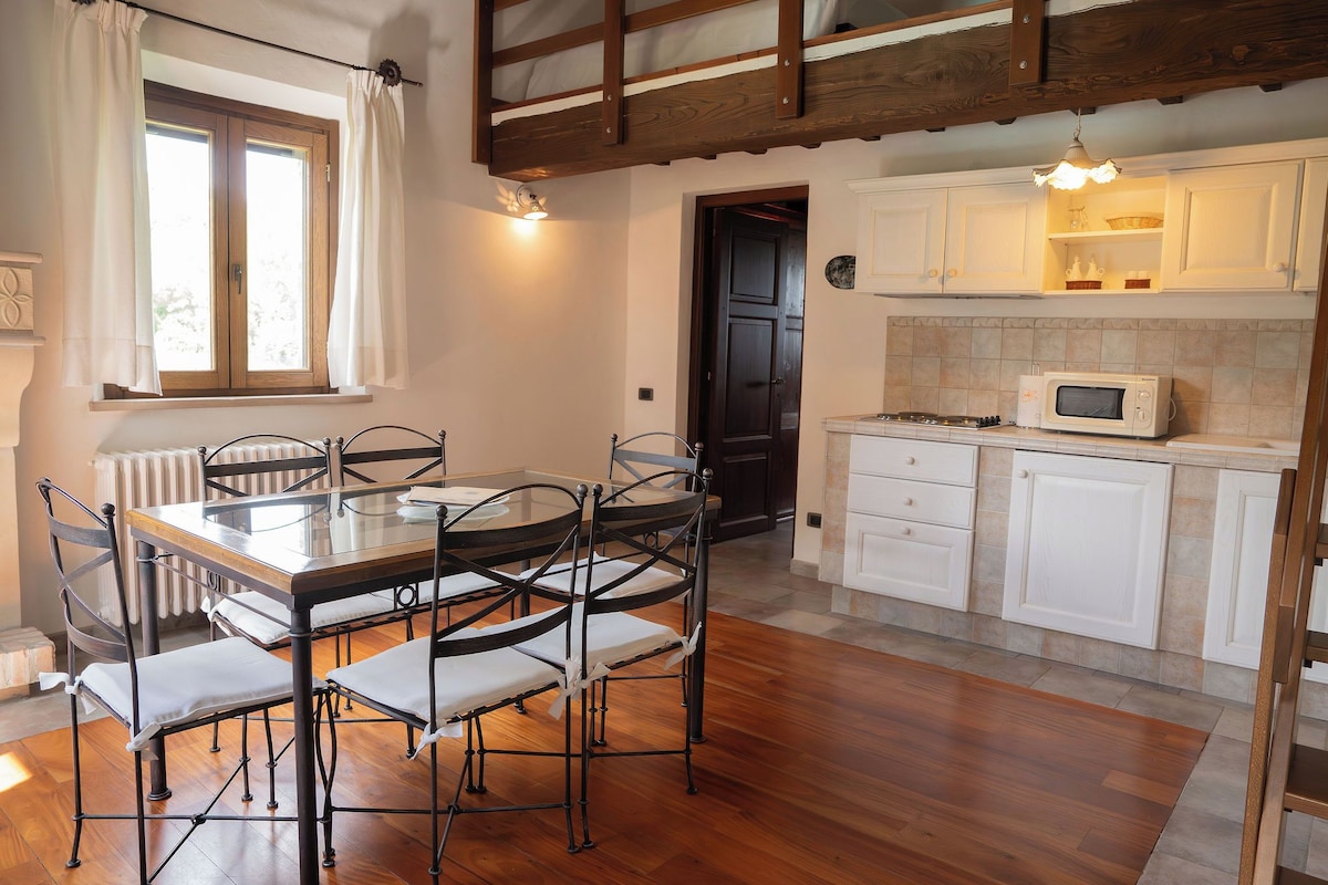 A dining area with a wooden table and chairs, a window with curtains, a kitchen with white cabinets and a microwave, and a wooden ceiling.