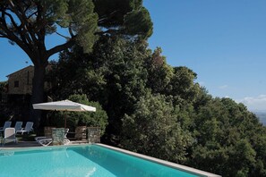 Une piscine entourée d’arbres et d’un bâtiment en pierre, avec des chaises longues et un parasol à proximité.