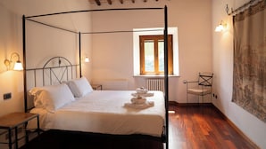Un letto a baldacchino con lenzuola bianche, pavimento in legno, una finestra con vista sul verde e un arazzo appeso al muro.