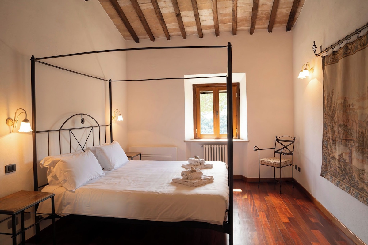 A four-poster bed with white linens, a wooden floor, a window with a view of greenery, and a tapestry on the wall.