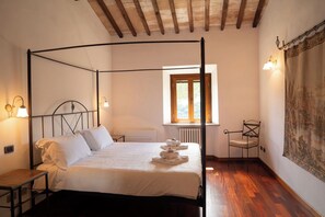 A four-poster bed with white linens, a wooden floor, a window with a view of greenery, and a tapestry on the wall. - Agriturismo Aia Leccioni (Pomarance)