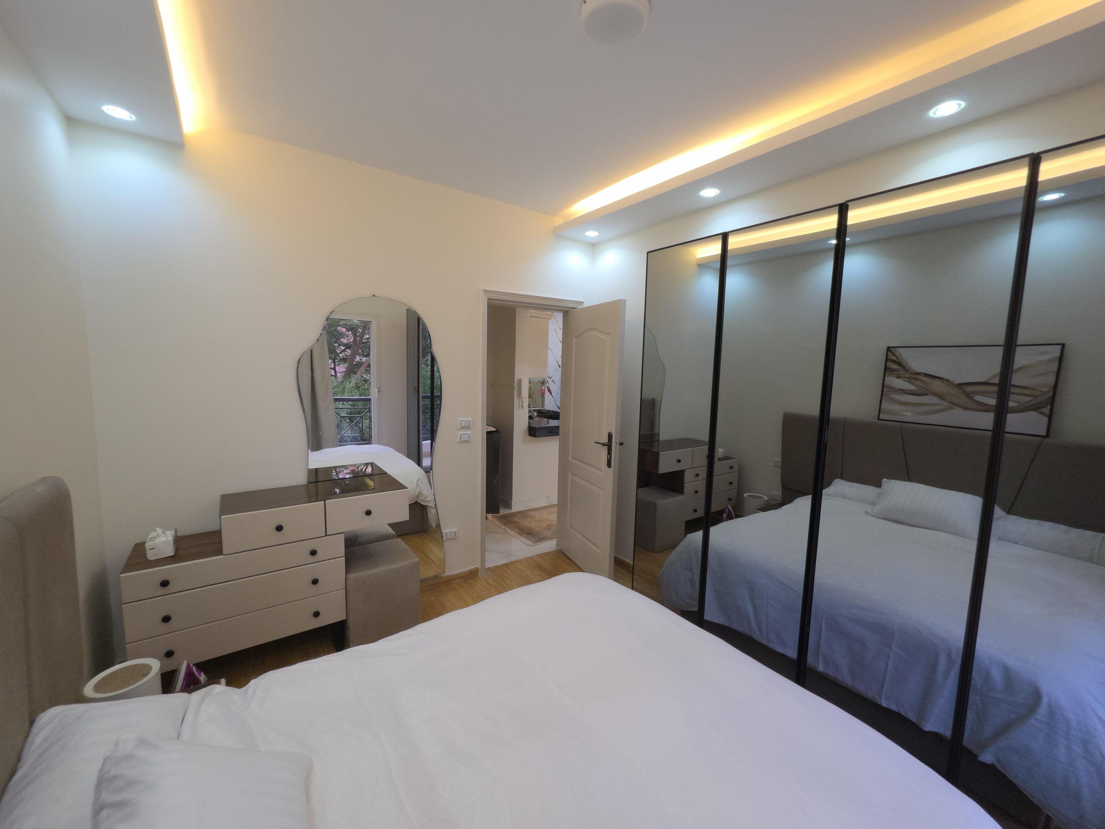 Une chambre moderne avec un grand lit, une commode, une coiffeuse avec un miroir et un dressing avec des portes coulissantes.