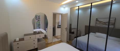 A modern bedroom with a large bed, a dresser, a vanity with a mirror, and a walk-in closet with sliding doors.