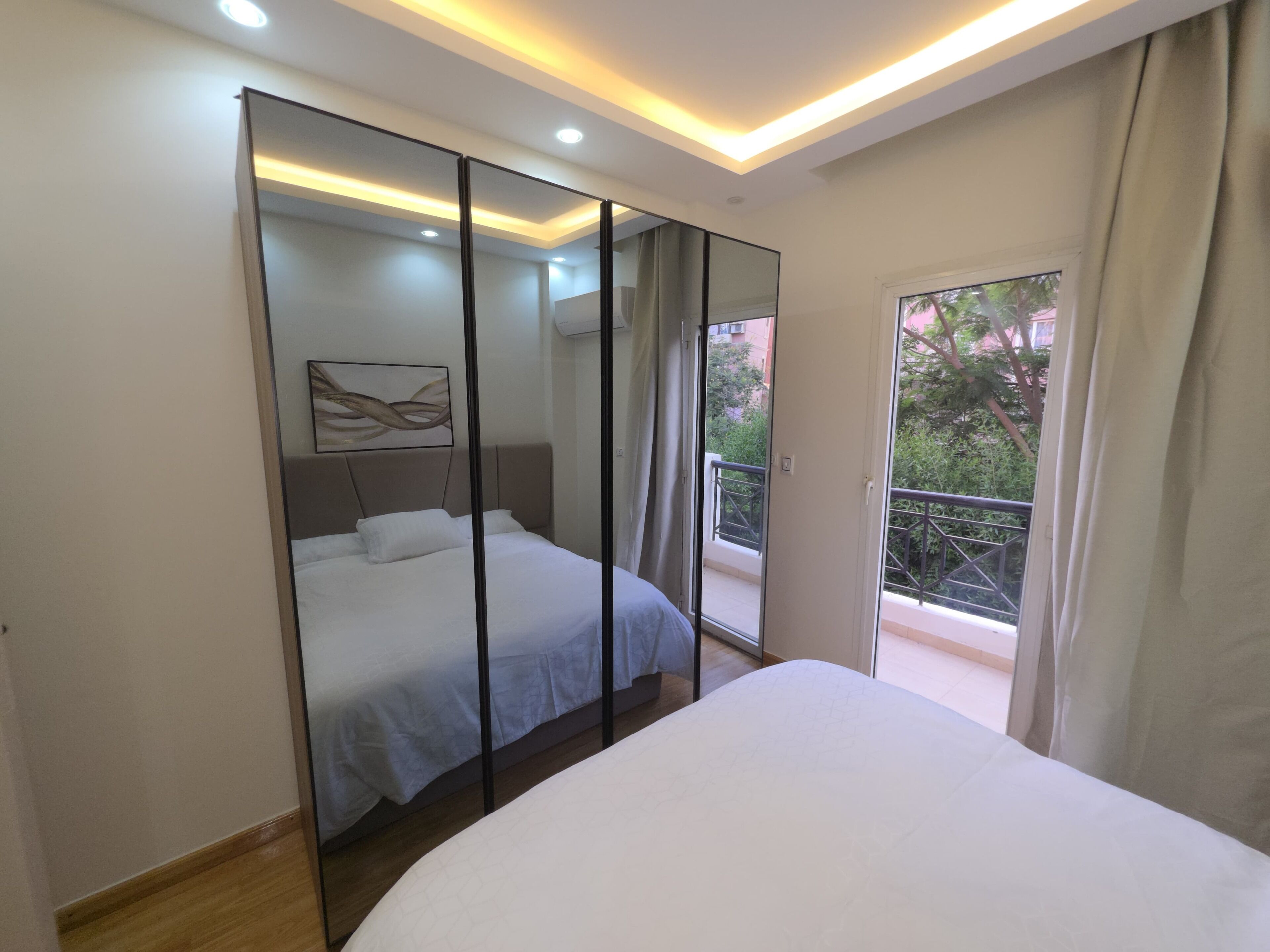 A bedroom with a bed, a large mirror, and a balcony with a view of greenery.