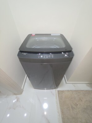 A compact, modern washing machine with a digital control panel, placed in a corner of a room with a white tiled floor. - Luxury stay in Sheikh Zayed, beside schools, malls (6th October)