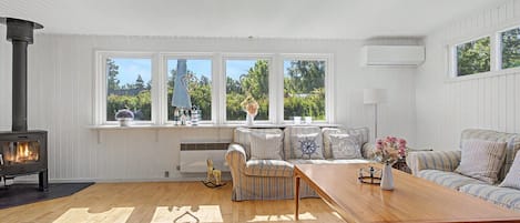 A cozy living room with a wood-burning stove, a sofa set, a wooden coffee table, and large windows offering a view of greenery.