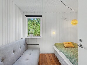 Ein kleiner, kompakter Raum mit einem grauen Sofa, einem Einzelbett mit einem grün gemusterten Bettbezug, einem Fenster mit Blick ins Grüne und einer Hängelampe.