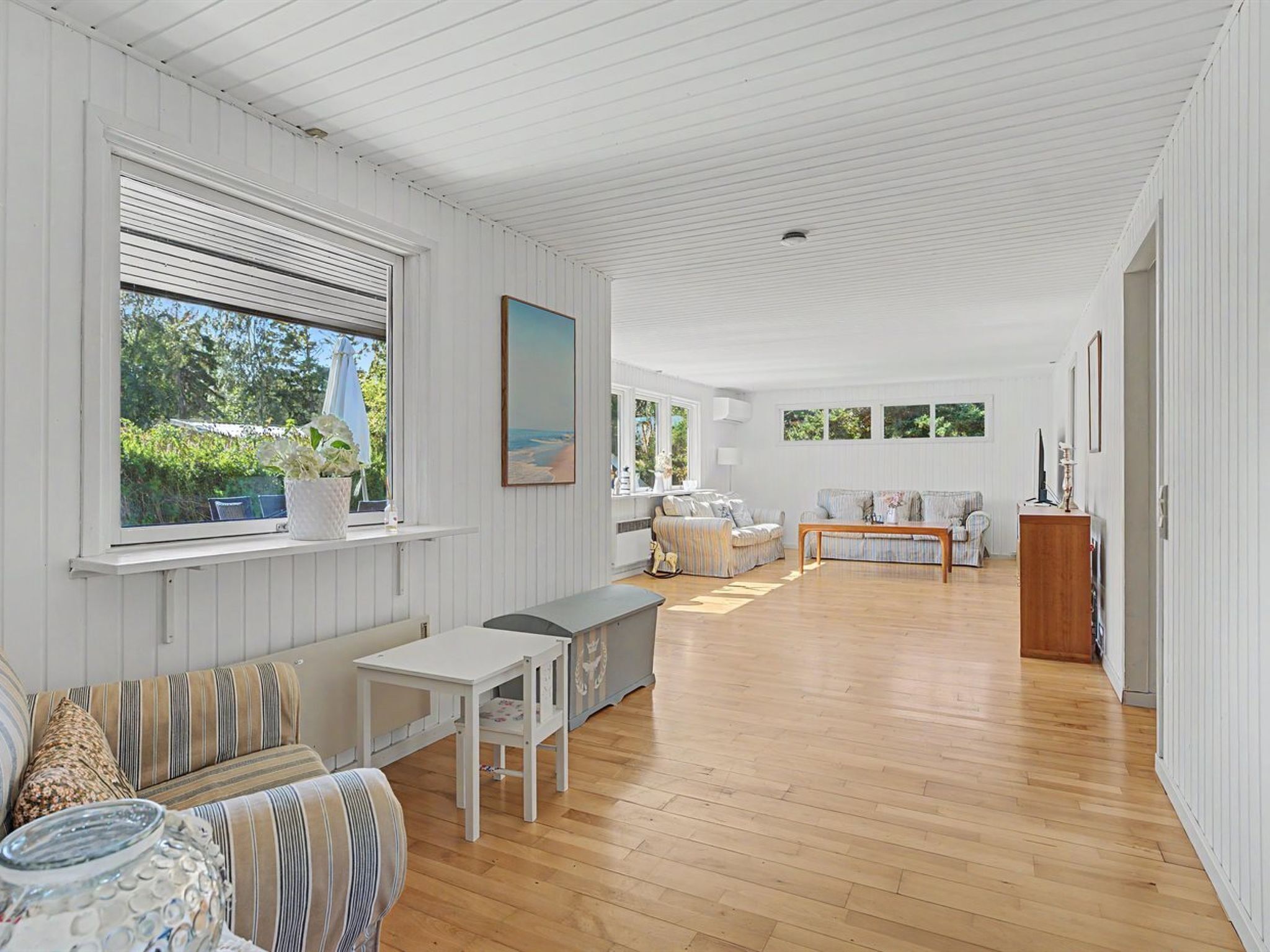 A spacious room with a wooden floor, white walls, and a large window overlooking greenery.