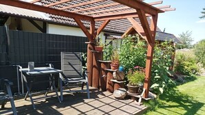 Een terras buiten met een houten pergola, een tafel en stoelen.